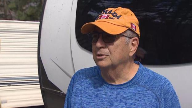 Man in blue shirt, yellow hat, and sunglasses standing in front of a white RV.